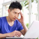 Worried Young Chinese Man Sitting At Desk Using Laptop At Home