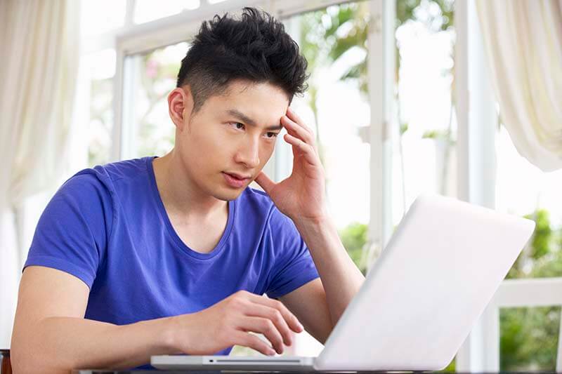 Worried Young Chinese Man Sitting At Desk Using Laptop At Home
